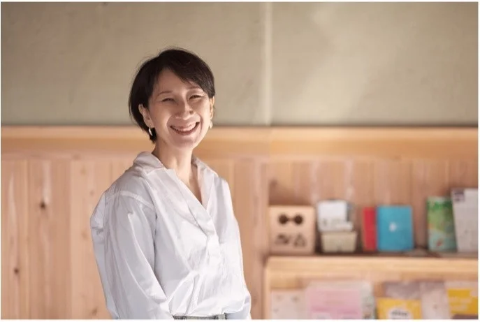 白いシャツを着たショートヘアの女性が、木製の棚を背景に明るく微笑んでいます。自然体で幸せそうな表情が印象的なポートレート写真です。