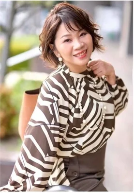 幾何学模様のブラウスとパールネックレスを身につけた笑顔の女性のポートレート。短めの髪型で、明るい表情を見せています。