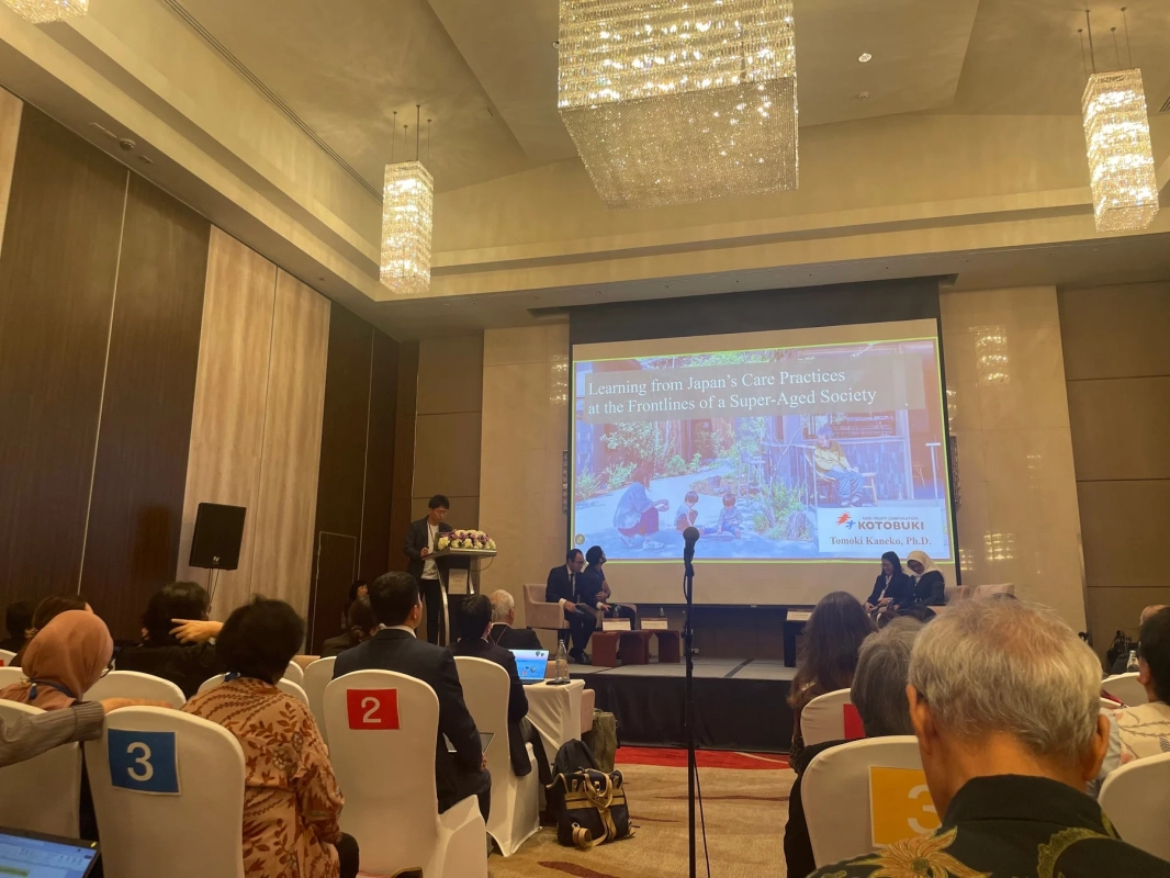 広い会場で行われている会議またはセミナーの様子です。演壇で発表者が講演しており、後方のスクリーンには「Learning from Japan's Care Practices at the Frontlines of a Super-Aged Society」というタイトルのスライドが映し出されています。会場には多くの聴衆が着席しています。