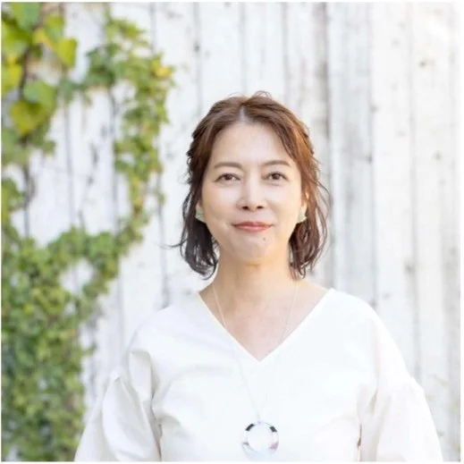 白いブラウスとネックレスを身につけた、笑顔の日本人女性のポートレート。背景には白い板壁と蔦が写っており、屋外で撮影されたことが伺えます。