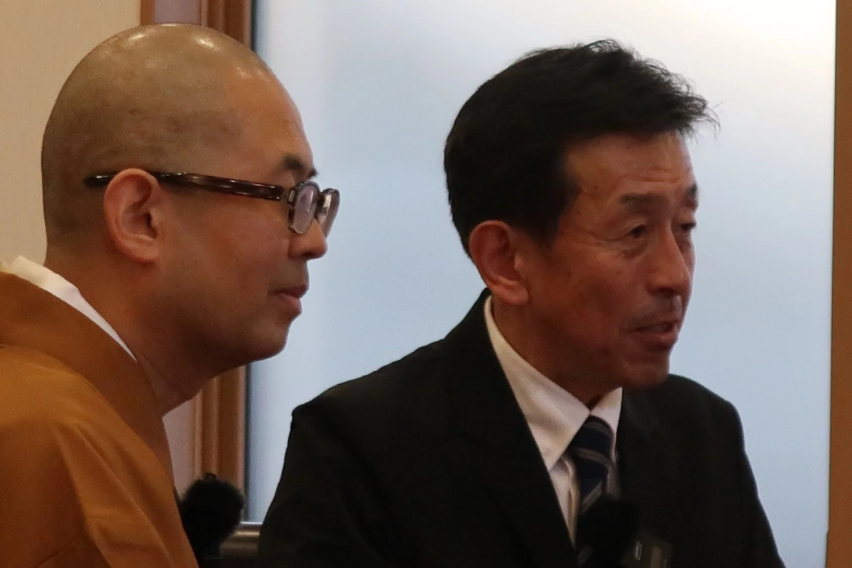 坊主頭の男性とスーツ姿の男性が並んで座っている