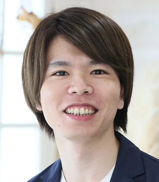 短い茶髪の若い男性が、紺色のジャケットを着用し、白い背景の前でカメラに向かって笑顔を見せているポートレート写真です。親しみやすい表情が印象的です。