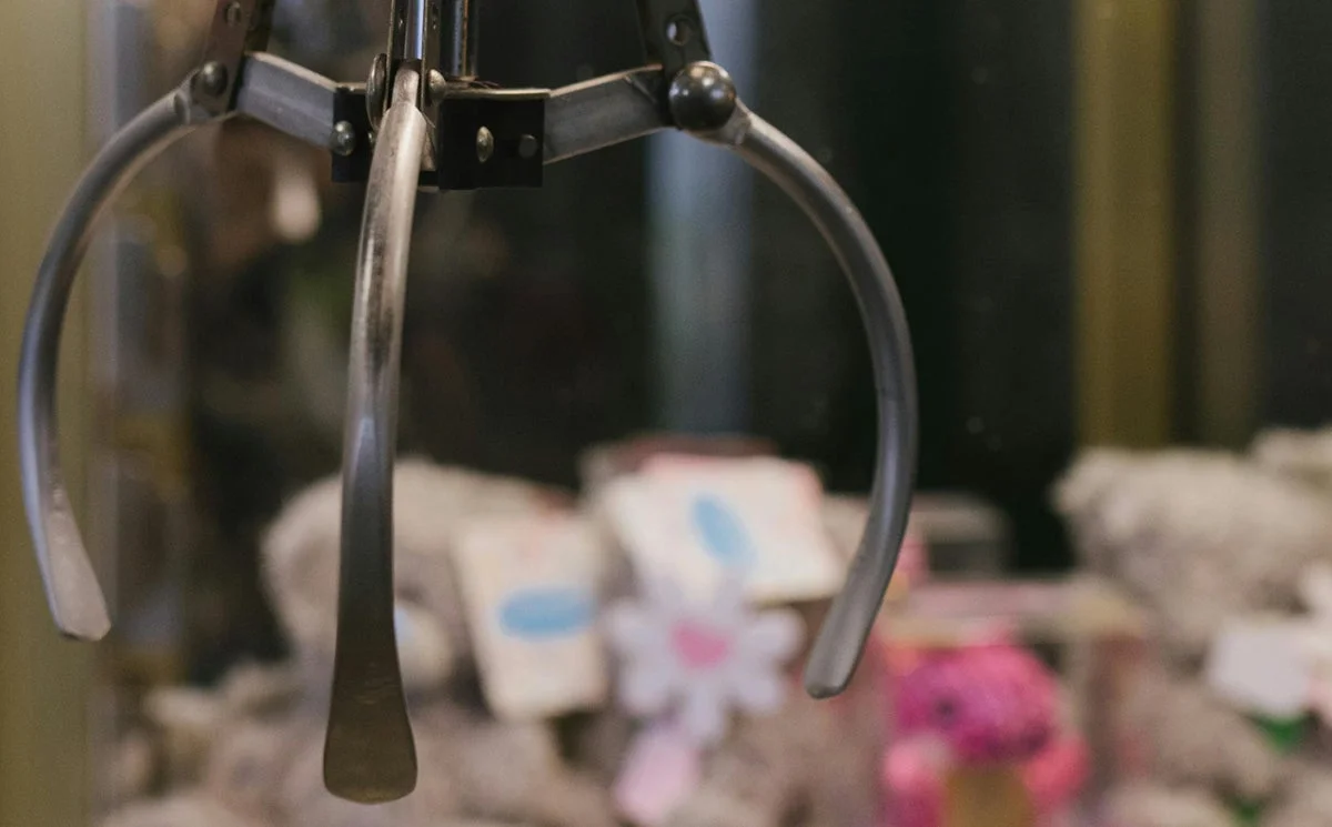 クレーンゲームのアームが、ゲーム機内の景品の上で待機している様子を捉えたクローズアップ画像です。背景にはぼやけたぬいぐるみなどの景品が見えます。