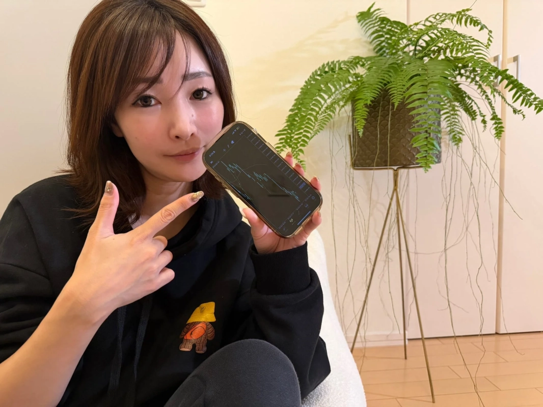 黒いパーカーを着た女性が、株価チャートが表示されたスマートフォンを見せています。