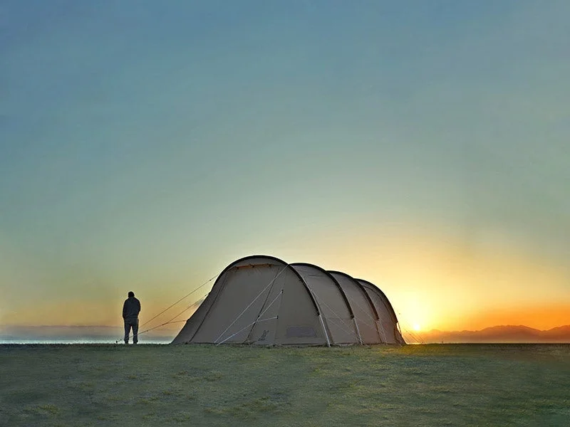 広大な草原に張られたテントのそばで、一人の人物が昇る朝日を眺めている風景