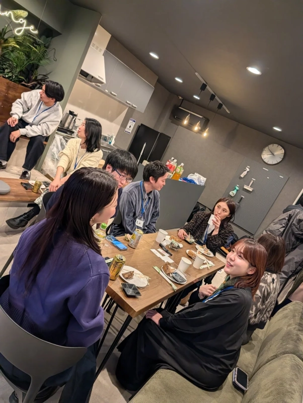 職場のラウンジで、複数の人々がカジュアルな雰囲気の中で食事や飲み物を囲み、談笑している様子