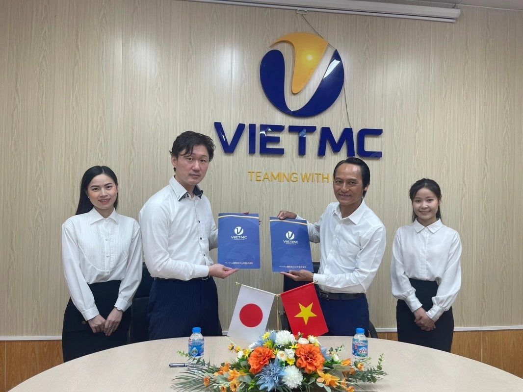 VIETMCのロゴがあるオフィスで、日本とベトナムの国旗が置かれたテーブルを囲み、男性2名と女性2名がビジネス契約書と思われる青いファイルを手にしている。