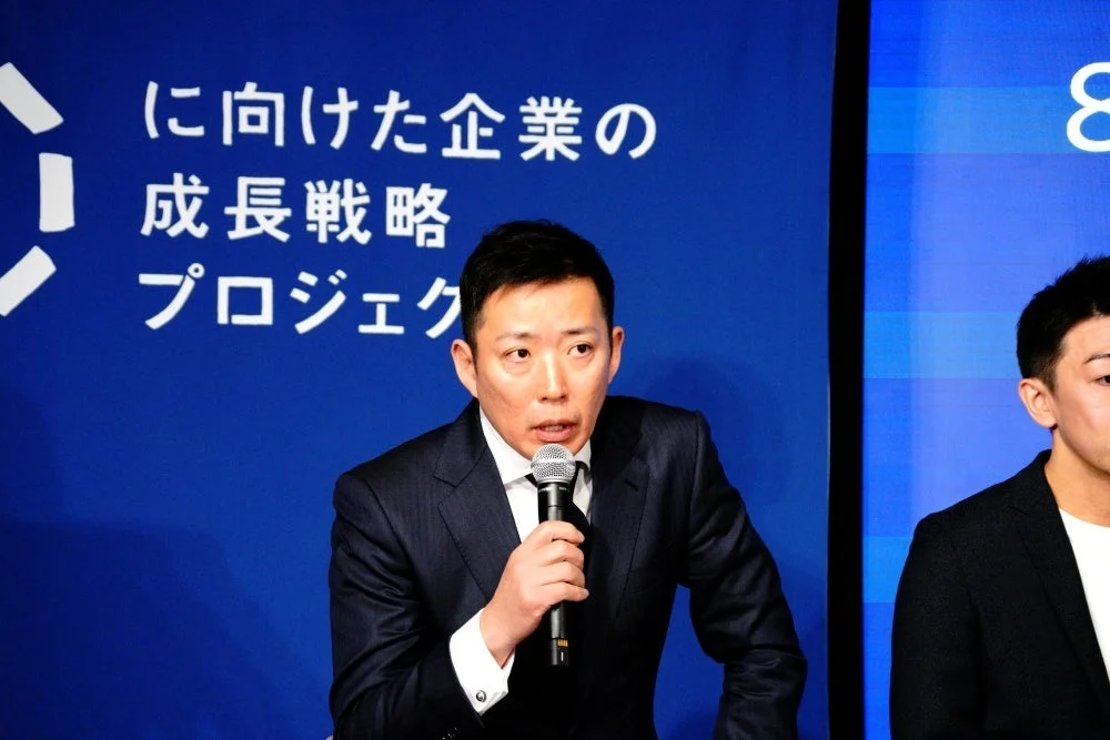 青い背景の前でスーツ姿の男性がマイクを持って話しており、企業の成長戦略に関するプレゼンテーションか会議の様子