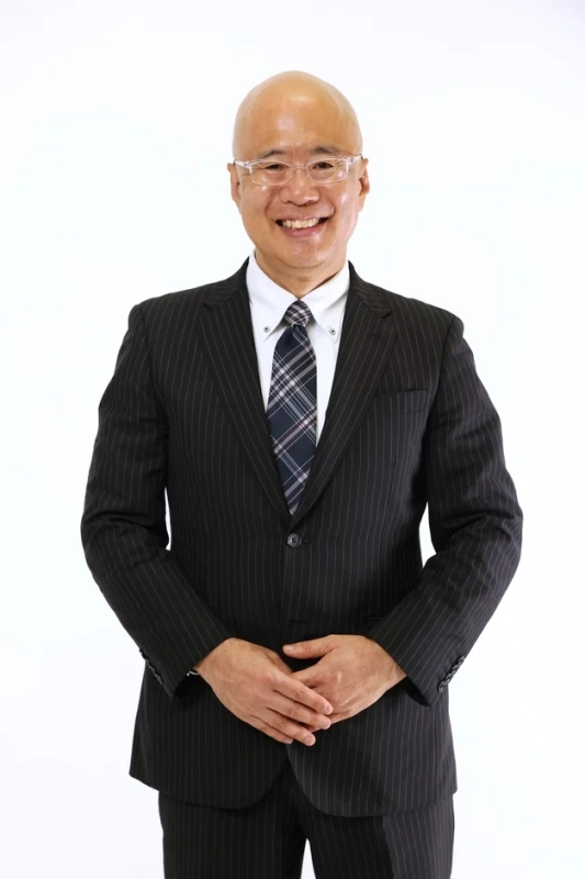 白背景の前で笑顔を見せる坊主頭の男性のポートレート。黒のストライプスーツにチェック柄のネクタイ、眼鏡を着用し、両手を組んで立っています。