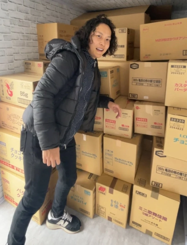 たくさんの段ボール箱が積み上げられた部屋で、黒いダウンジャケットを着た男性がカメラに向かって微笑んでいます。段ボール箱にはお菓子などの商品名が書かれており、倉庫やストックルームのような雰囲気です。