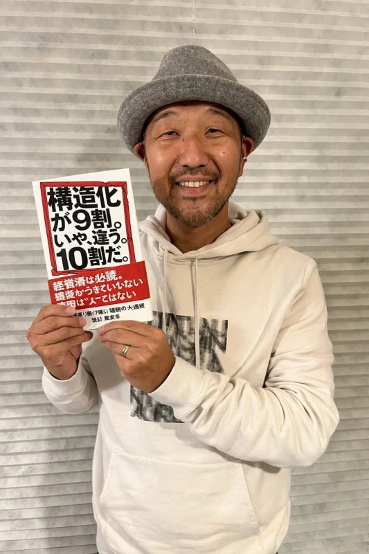 グレーのハットと白いパーカーを着た男性が、笑顔で「構造化が9割。いや、違う。10割だ。」というビジネス書を持っている。