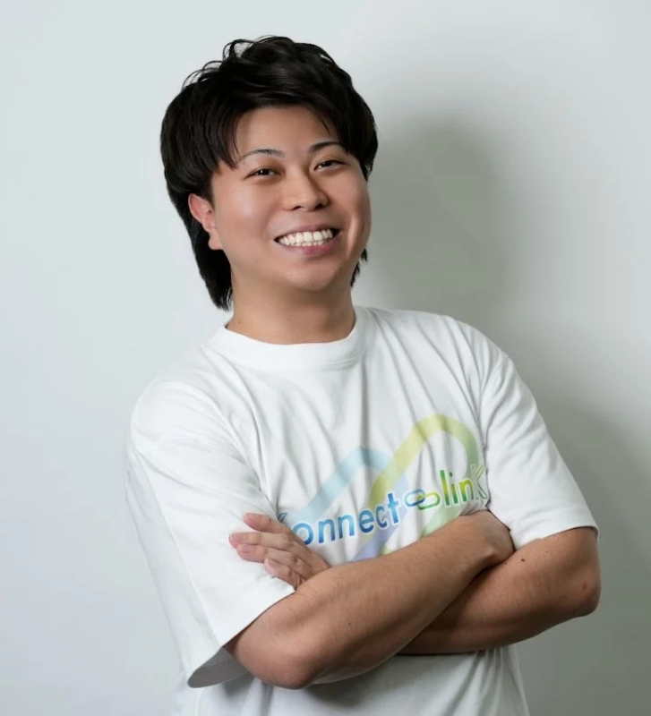 白いTシャツを着た若い男性が腕を組み、カメラに向かって満面の笑みを浮かべているポートレート写真