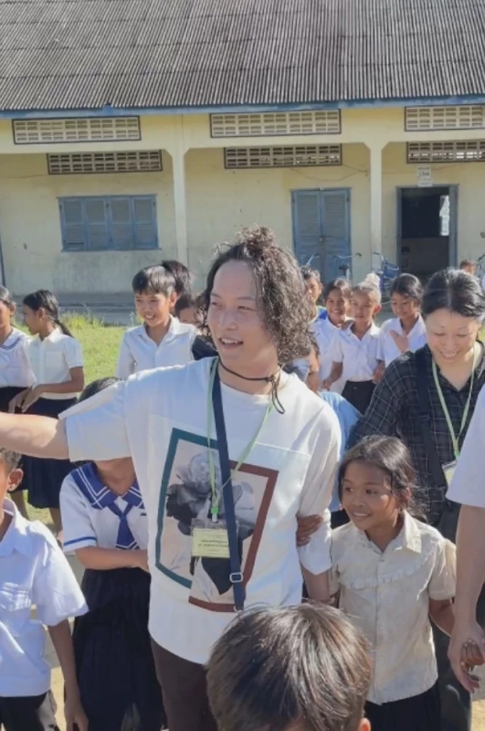 屋外で多くの子どもたちと大人たちが笑顔で交流している様子を捉えた写真です。白いTシャツの男性が中心に立ち、周囲の子供たちも楽しそうです。背景には学校のような建物が見えます。