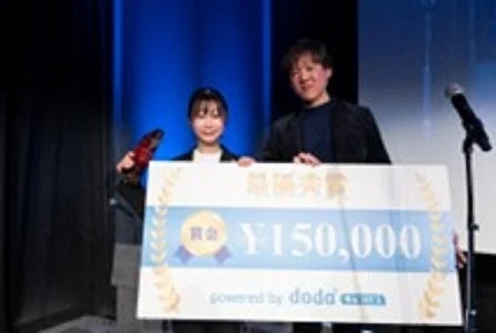 ステージ上で男女2人が最優秀賞の賞金15万円が書かれた巨大な小切手を受け取っています。dodaが協賛するイベントでの表彰式の様子です。