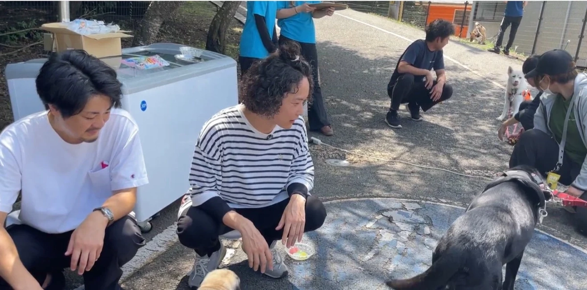 屋外で数人の人々が犬たちと交流している様子です。白いフリーザーが置かれており、何らかのイベントや集まりが行われているようです。リラックスした和やかな雰囲気が感じられます。