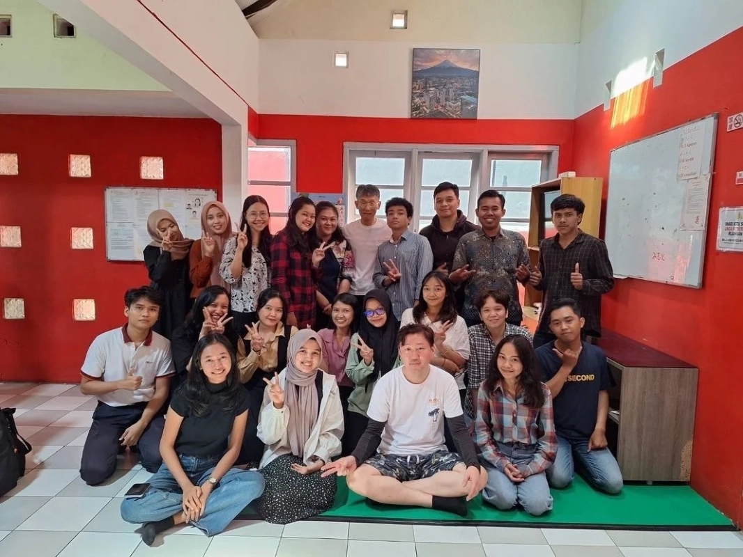 赤い壁の部屋で、大勢の若者が笑顔でピースサインをしながら集合写真を撮っている。