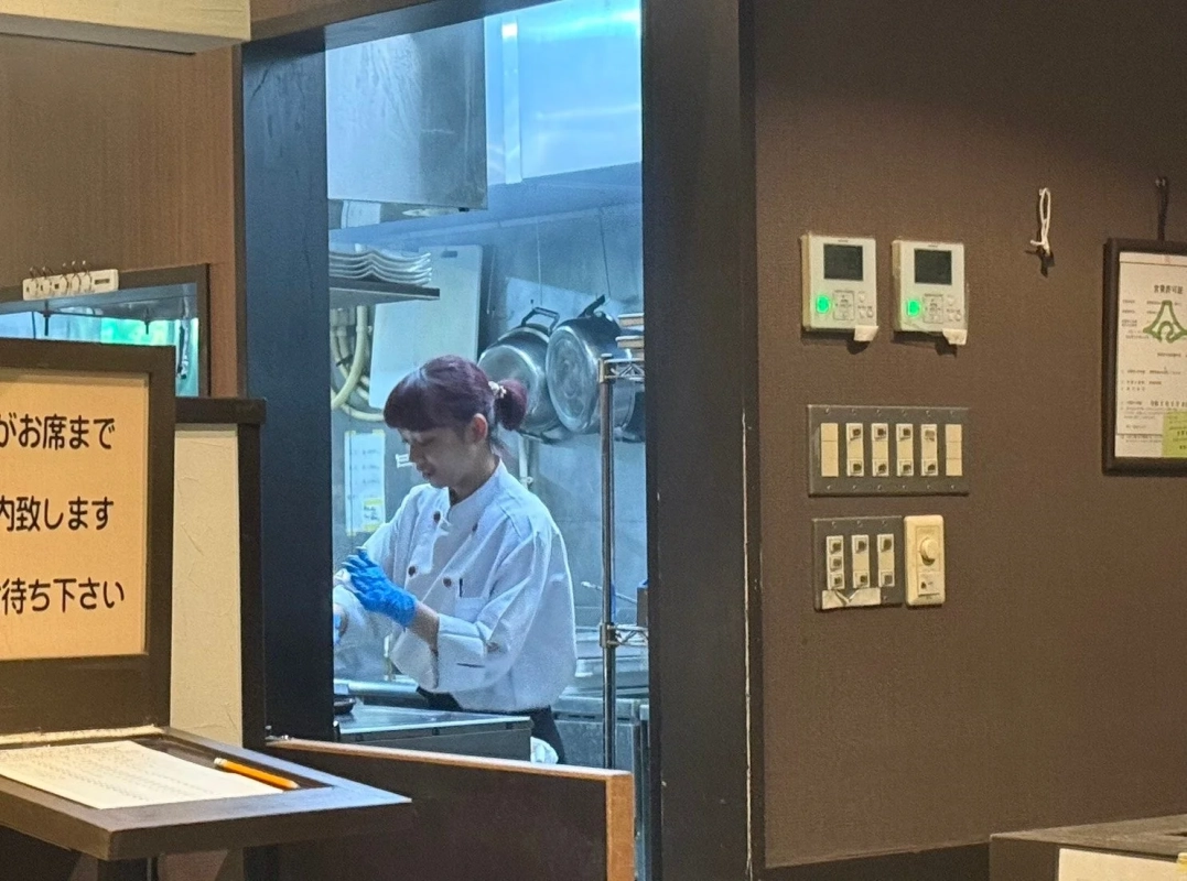 白いコックコートと青い手袋を着用した女性料理人が厨房で調理作業をしている様子。