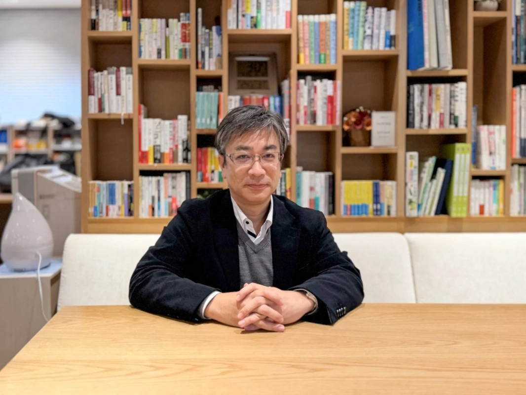 眼鏡をかけた中年の日本人男性が、本棚を背景にした木製のテーブルに座り、カメラの方を見ています。落ち着いた雰囲気のビジネスシーンです。