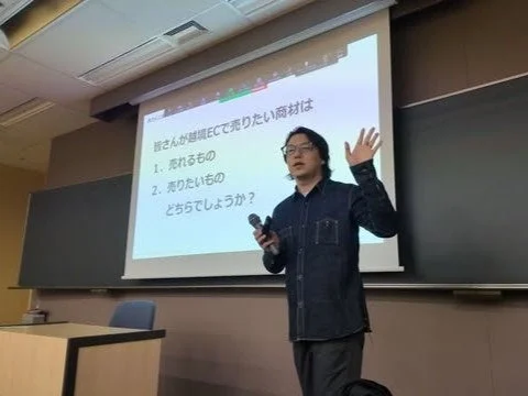教室のような空間で、一人の男性がプロジェクタースクリーンを背景にプレゼンテーションを行っています