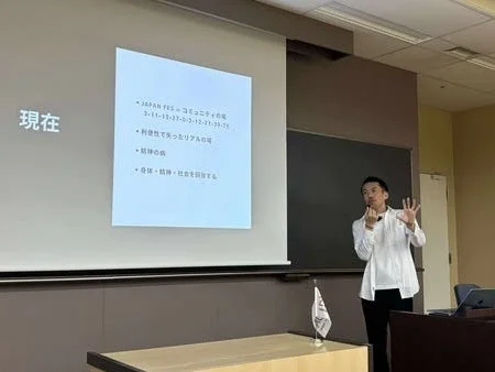 会議室のような場所で、一人の男性がプロジェクターに映し出された日本語のスライドを背景にプレゼンテーションを行っています