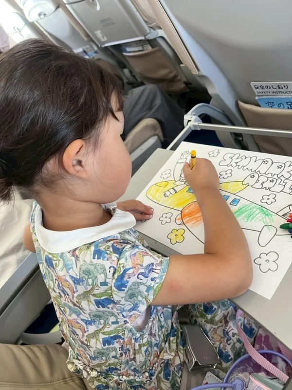 飛行機の座席で小さな女の子がクレヨンを使って塗り絵を楽しんでいます。