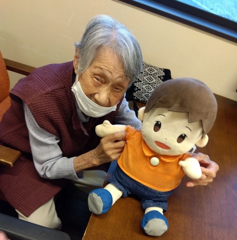 マスクを着けた高齢の女性が、男の子のぬいぐるみと一緒に満面の笑みを浮かべている