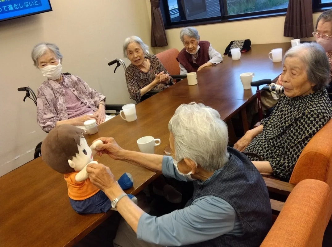 高齢者施設で、数人のお年寄りが大きなテーブルを囲んで座っており、そのうちの一人がテーブルに置かれた男の子のぬいぐるみと優しく触れ合っている