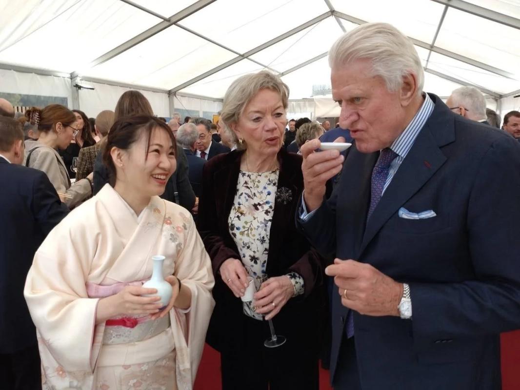 着物姿の日本人女性が笑顔で、小さな器から何かを試飲している外国人男性と交流している様子