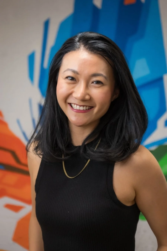 黒いノースリーブを着て金色のネックレスをつけたアジア人女性が、カラフルな抽象的な背景の前で笑顔を見せているポートレート写真