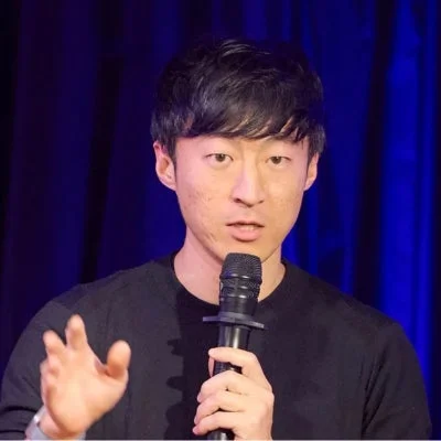 マイクを持ち、左手を上げて講演しているアジア人男性