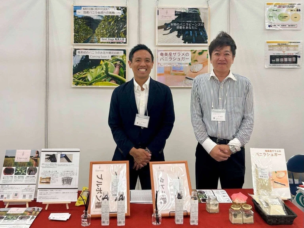 展示会ブースで2人の男性が、国産バニラ栽培やインドネシア産バニラビーンズ、奄美産ザラメのバニラシュガーといった「Eternal」ブランドのバニラ関連商品を展示・紹介しています