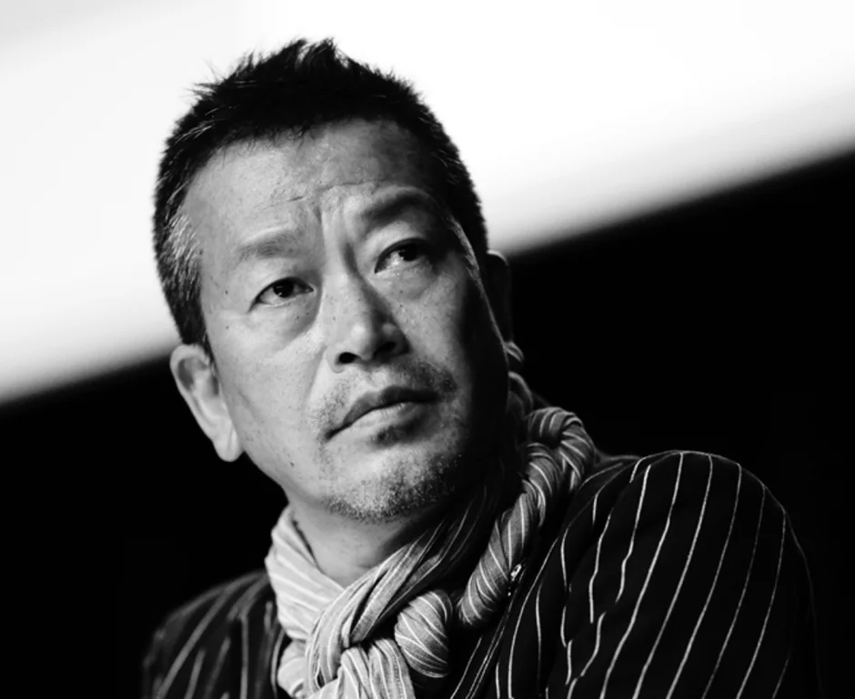 白黒写真に写る、ストライプのジャケットとスカーフを着用した中年のアジア人男性のポートレート。彼は真剣な眼差しで、何かを思案しているかのように斜め上を見つめている。