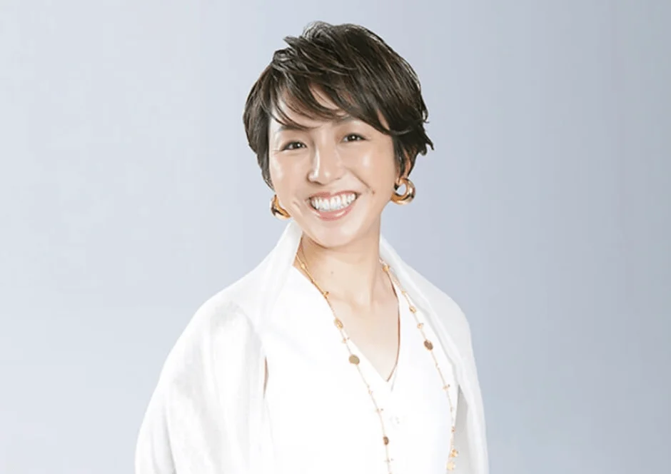 白い衣装とゴールドのアクセサリーを身につけたショートヘアの女性が、明るい笑顔を見せています。清潔感のあるポートレート写真です。