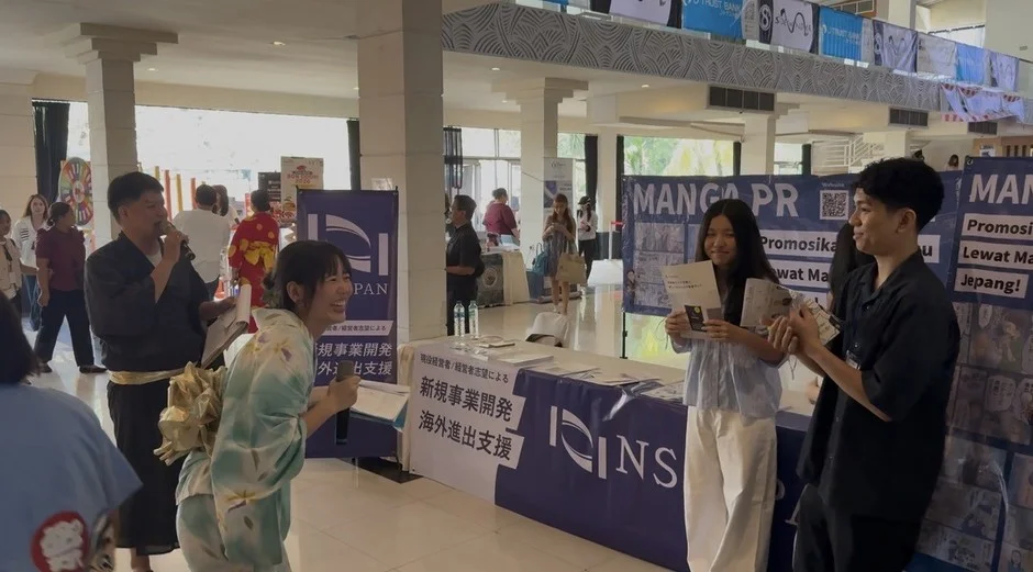 日本の漫画やビジネス支援に関するイベント会場の様子です。和服を着た人々がブースに集まり、一人の男性がマイクで話しています。新規事業開発や海外進出支援のバナーも見え、国際的な交流の場であることが伺えます。